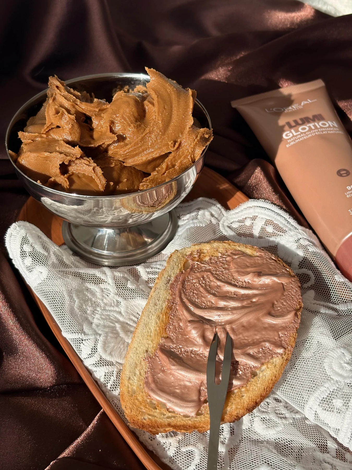 L’Oréal Paris Lumi Glotion styled beside toast and creamy textures on chocolate satin and lace